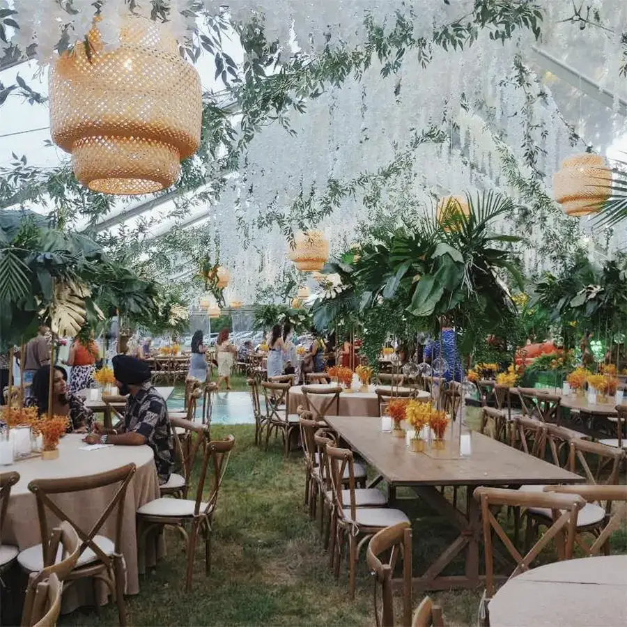 Tropical themed wedding with rattan chandelier lighting, lush greenery, and cross-back chairs under a clear top tent